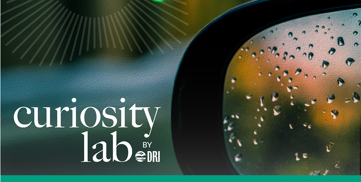 Curiosity Lab by DRI sunburst logo over an image of a side view mirror on a car reflecting the background.