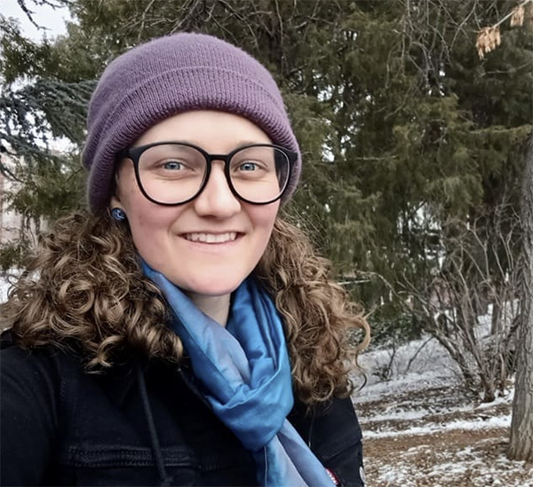 Selfie style photo of Abigail outside with pine trees in winter wearing a knit hat, scarf, and coat.
