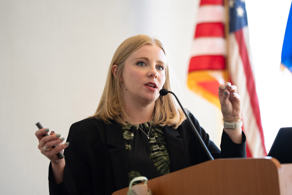 Ashley presenting at a podium with a microphone and American flag in the background.