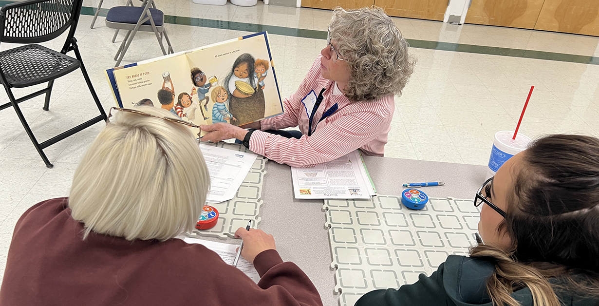 An instructor reads from a book to the educators at the table with robots in front of them. 