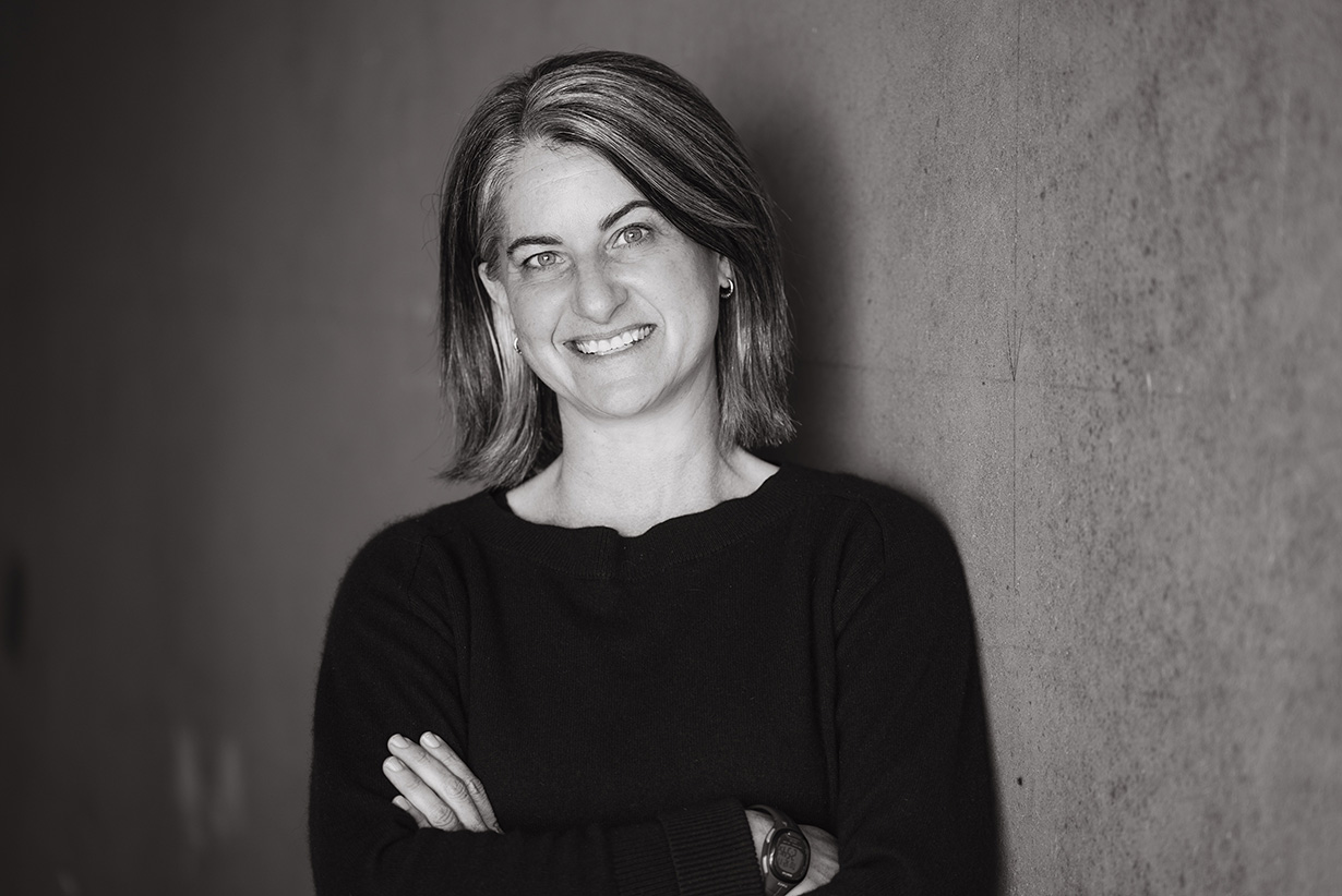Black and white headshot of Cayenne leaning against a wall with arms folded and smiling.