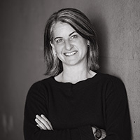 Black and white headshot of Cayenne leaning against a wall with arms folded and smiling.