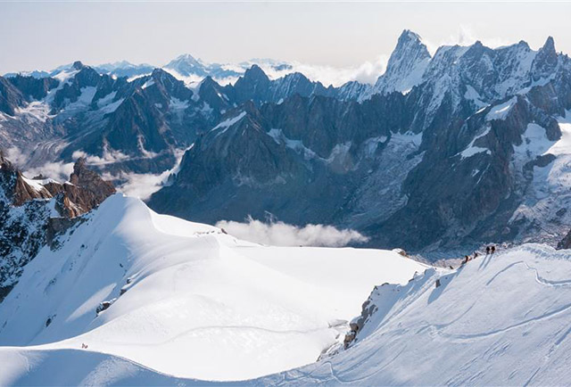 View of rocky and snow covered mountain peaks with hikers on a ridge. 