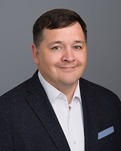 Headshot of Cleat smiling and wearing a suit in front of a grey background. 