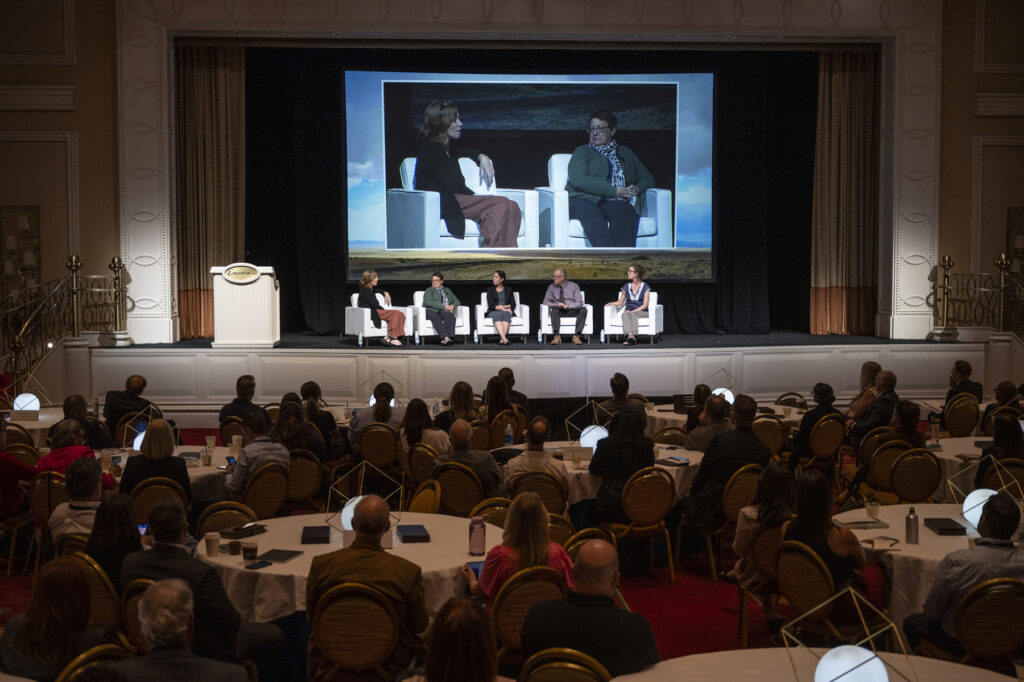 Darkened conference room with panelists on stage sitting in chairs. The audience is seated at round tables.