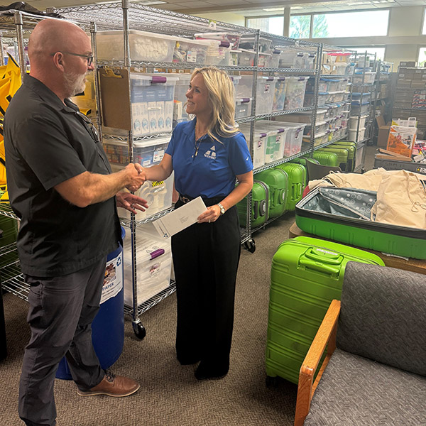 Two people are looking at each other and shaking hands in front of shelves of supplies and green boxes.