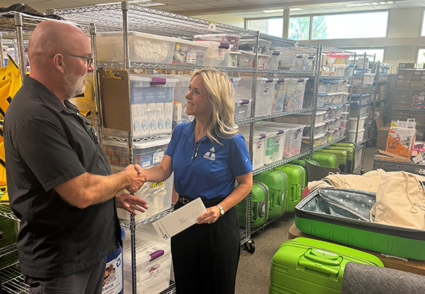 Two people are looking at each other and shaking hands in front of shelves of supplies and green boxes.