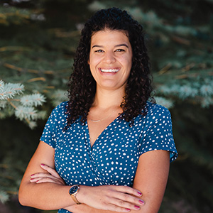 Emily McD-Williams Our Team 300px Headshot of Emily outside in front of pine trees smiling and wearing a blue shirt.