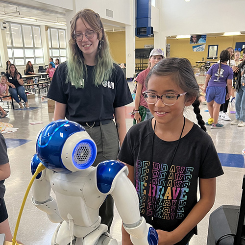 FR-Student-smiling-at-robot Student smiling at a blue and white robot with a teacher looking on.