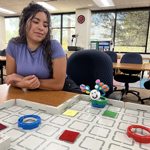 FR metrics grid table with robot An educator looks over robots on a grid table with obstacles that form a pathway.