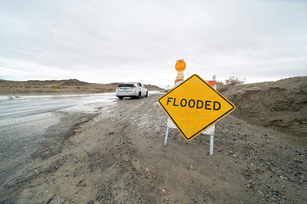 A yellow sign that says Flooded is set at the side of a road with a vehicle parked on the washed out shoulder.
