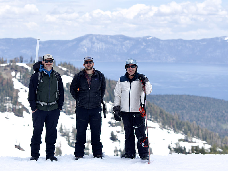 Three men in winter gear and snow board on a peak overlooking the water of Lake Tahoe.