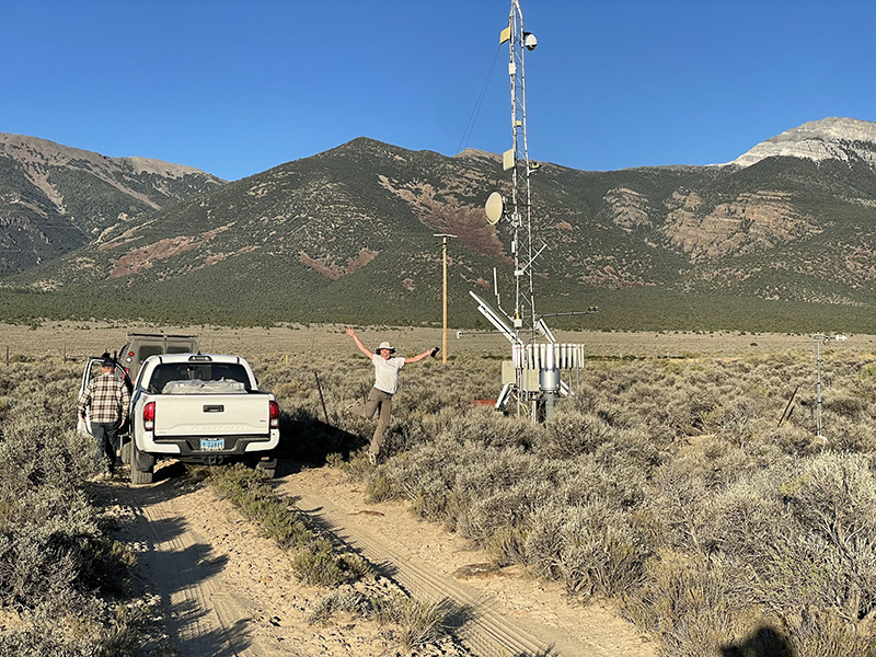 A researcher jumping with their arms stretched out and smiling in a landscape view of a weather station in the valley with mountains in the background.