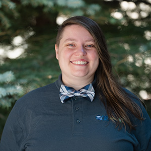 Ileah Kirchoff Our Team 300px Headshot of Ileah in front of pine trees and smiling wearing a DRI blue collared shirt with a bow tie.
