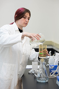 A researcher in a lab wearing a white lab coat pouring liquid into a beaker. 