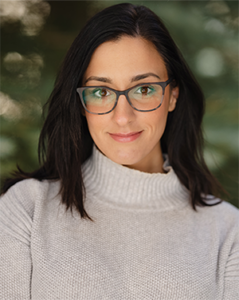 Headshot of Dr. Kathleen Rodrigues wearing a sweater and glasses against a background of pine trees. 