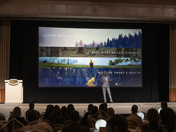Kumud Acharya on Stage AWE Kumud Acharya speaking on the stage at AWE+ Summit 2024 with images of wildfire behind him.