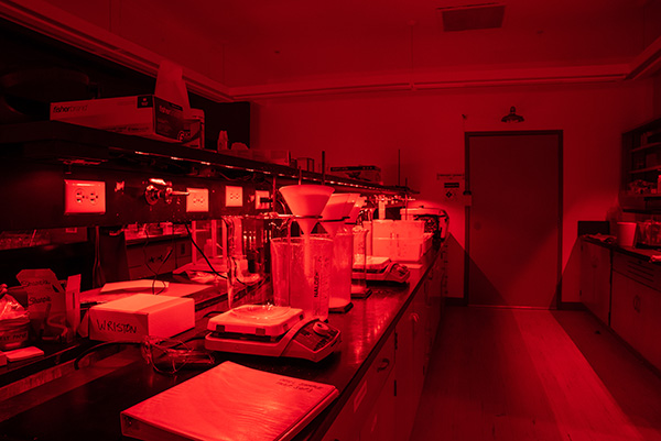Luminescence-005_Web A close up of a lab center island and equipment on the counter bathed in a deep red glow.