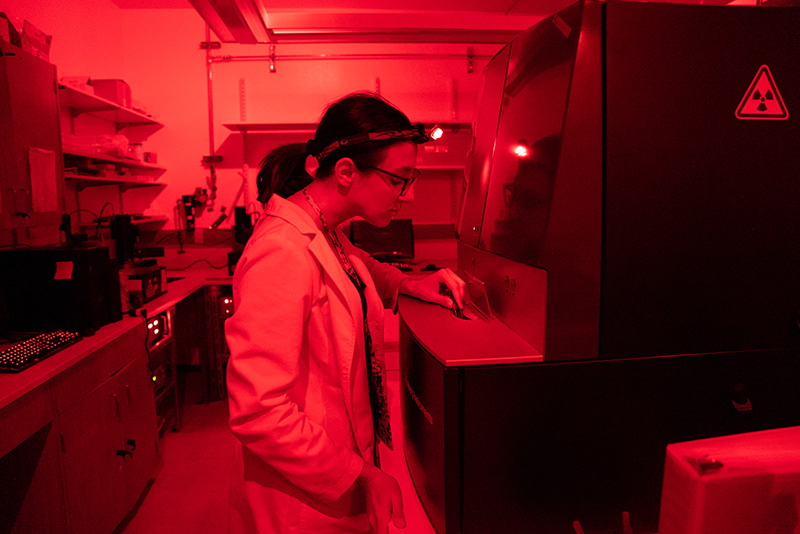 Luminescence-107_Web A researcher works with a piece of large equipment in a lab with a red glow.