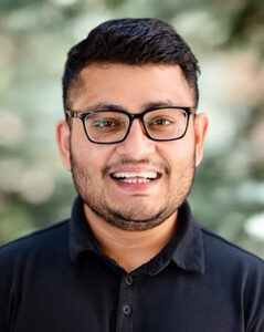 Headshot of Monty Majumdar against a background of pine trees wearing glasses and smiling. 