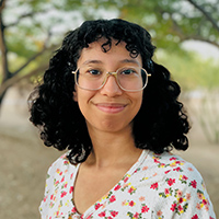 Photo of Myriam outside in front of trees in a flowered shirt and wearing glasses.