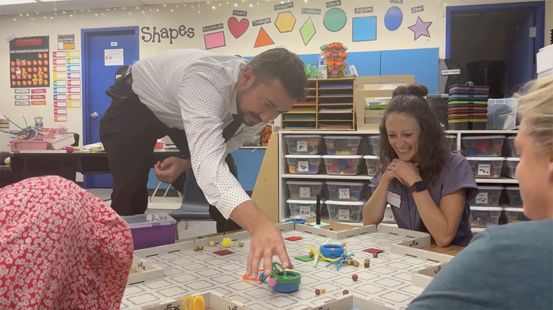 Impact Image An educator reaches for a robot on a grid with other educators look on smiling.