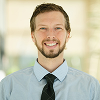Headshot of Patrick wearing a tie and collared shirt and smiling in a brightly lit hallway full of plants.