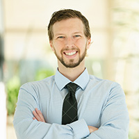 Headshot of Patrick Hurbain in a collared shirt and tie in a brightly lit hallway with plants. 