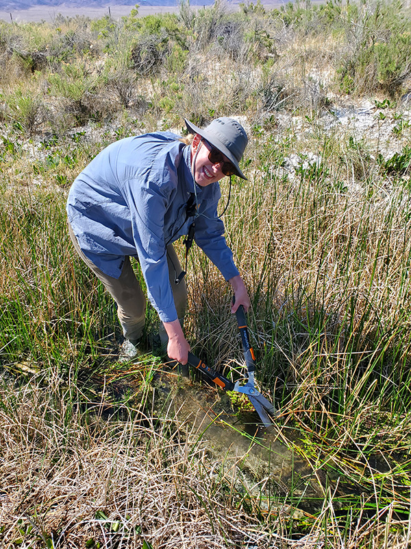 Alexa Daniel wearing a sunhat and sunglasses in a stream of water and cutting back grass.