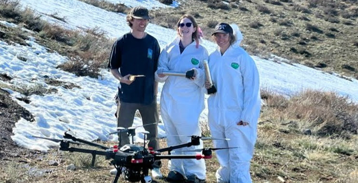 Three students stand on a hillside around a large drone. 