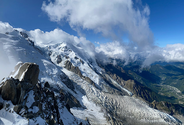 View of a snow covered side of a mountain with blue sky and clouds in the distance.
