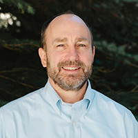 Headshot of Sean McKenna in a blue collared shirt and pine trees in the background.