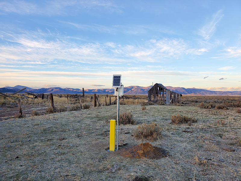 A meter on a pole in a desert landscape with a dilapidated barn and mountains in the distance.