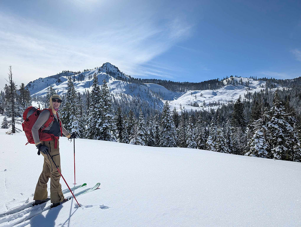 Alexa Daniel with skis and poles in a winter landscape with mountains and pine trees.