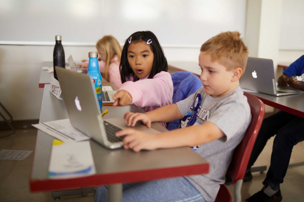 A child in a pink sweater pointing at a laptop screen with a boy sitting next to her.