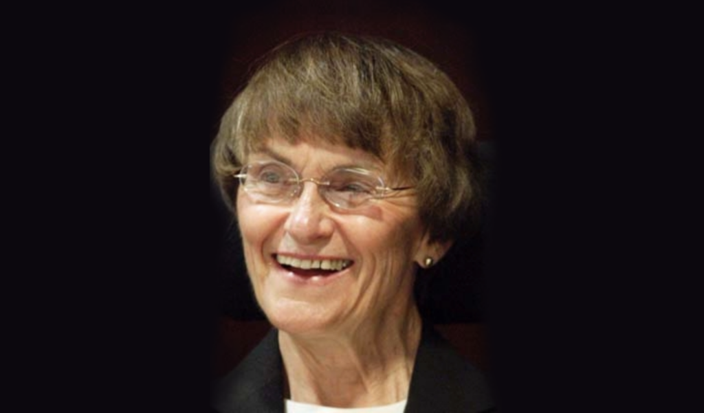 Sue Wagner wearing glasses and smiling at something just off camera against a black background.