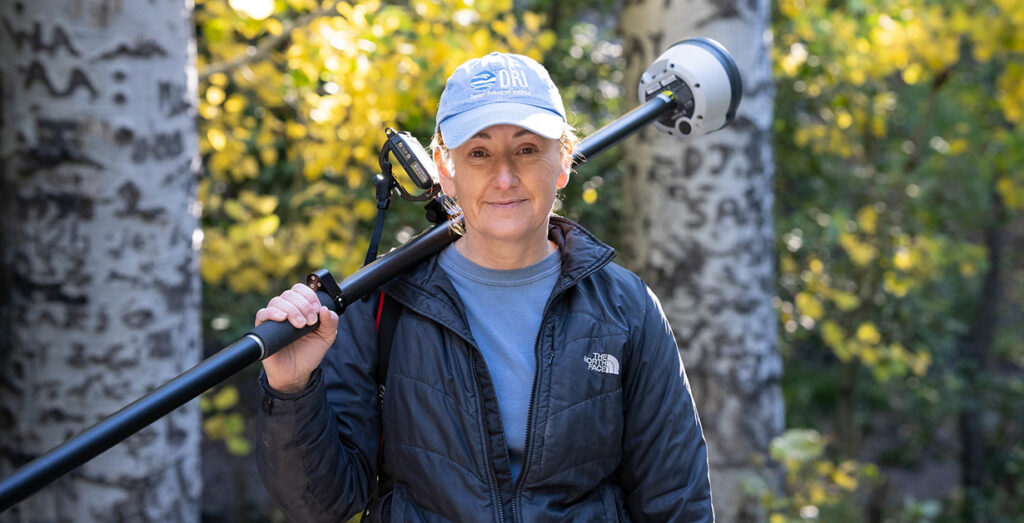 Teresa Wriston outside in front of trees holding a piece of equipment and wearing a DRI hat.