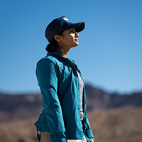 Tiffany is looking at the horizon in a desert landscape wearing a blue jacket and DRI hat. 