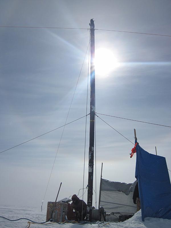 Tall ice core drill reaching up from the ice to the sun in the background.