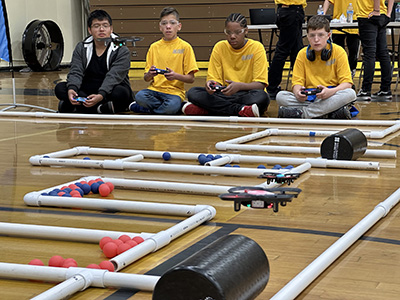 Website_Drone-1-scaled Students with controllers sitting on the edge of a track and flying drones through obstacles.