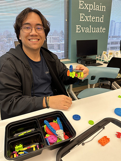 Website_Home-page-4-scaled An Educator smiling and holding up a small robot that they built with various gears at a training session.
