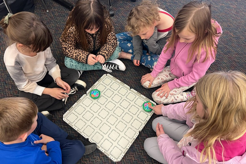 Six students gathered around a grid on the floor and controlling robots in the squares.